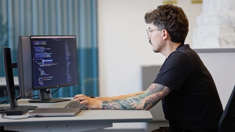 En programmør med tatoveringer sidder ved et skrivebord og arbejder på en computer med kode vist på skærmen.